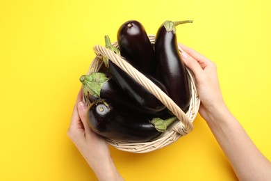 Photo of Woman with wicker basket of fresh eggplants on yellow background, top view