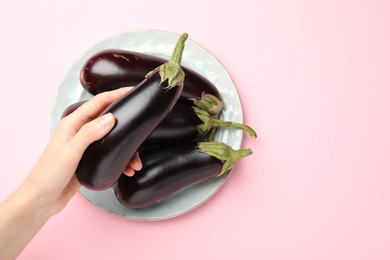 Photo of Woman holding fresh eggplant above plate with whole ones on pink background, top view. Space for text