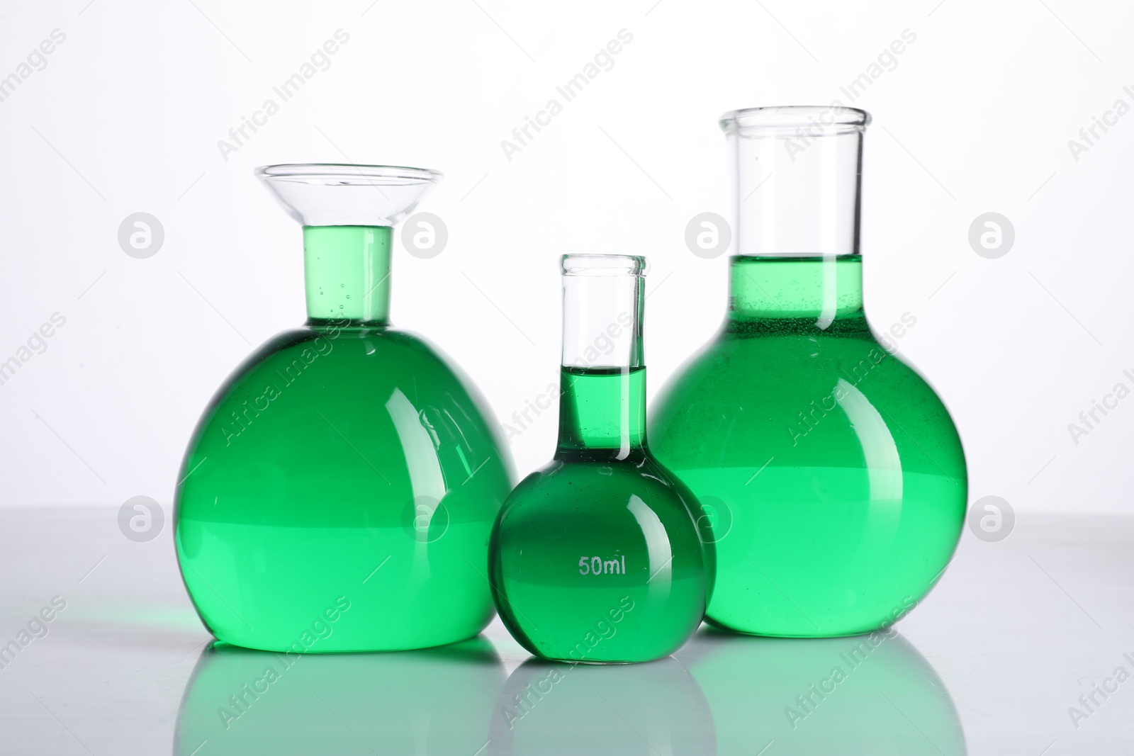 Photo of Different laboratory glassware with green liquid on mirror surface against white background