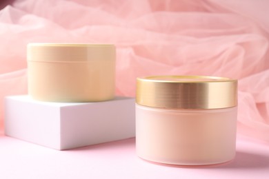 Photo of Promotion. Jars of cosmetic products, podium and tulle cloth on pink background, closeup