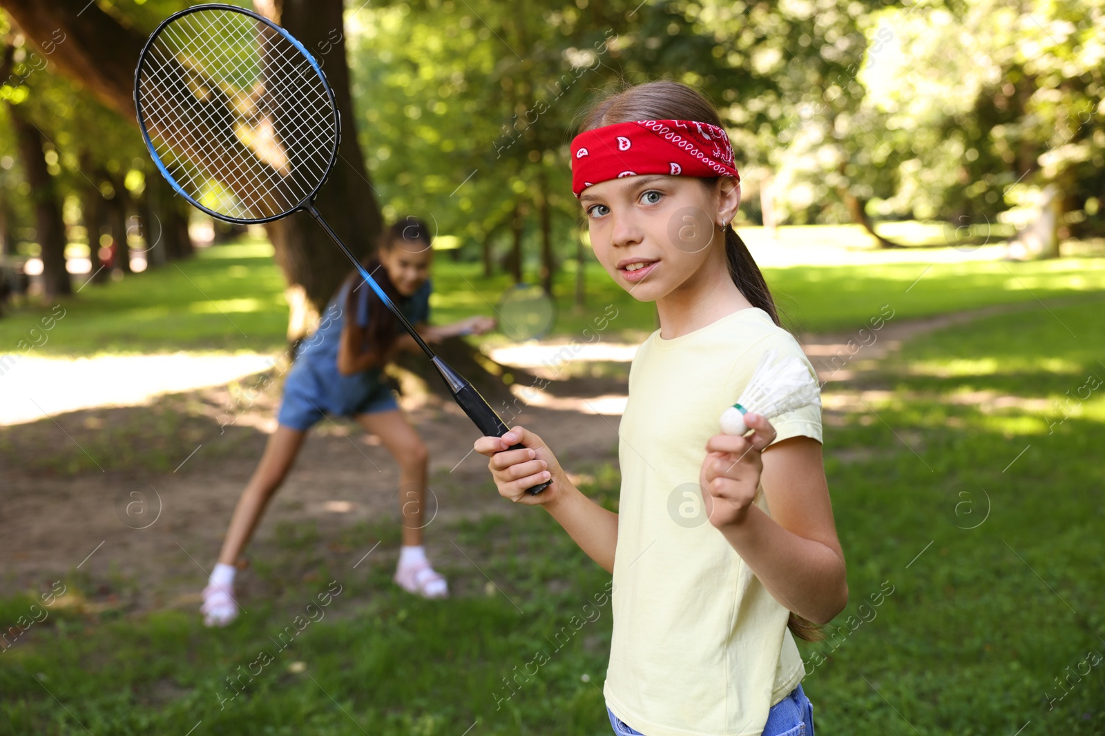 Cute little girl in bandana with badminton racket and shuttlecock playing with her friend in park, selective focus Photo of Cute little girl in bandana with badminton racket and shuttlecock playing with her friend in park, selective focus