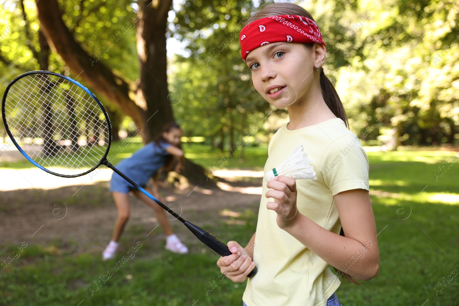 Smiling little girl in bandana with badminton racket and shuttlecock playing with her friend in park, selective focus Photo of Smiling little girl in bandana with badminton racket and shuttlecock playing with her friend in park, selective focus