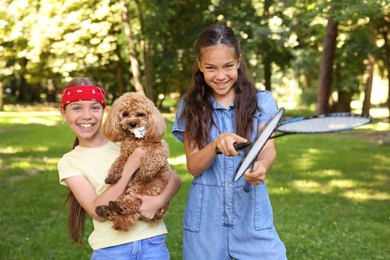 Photo of Smiling little girls with badminton rackets having fun with their dog in park