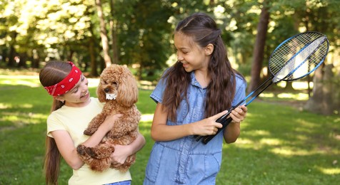 Photo of Smiling little girls with badminton rackets having fun with their dog in park