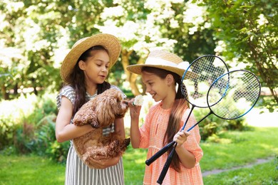 Photo of Smiling little girls in hats with badminton rackets and shuttlecock having fun with their dog in park