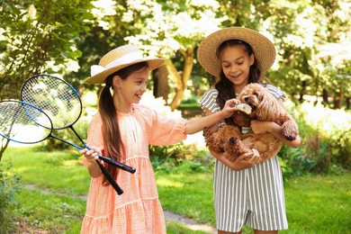 Photo of Smiling little girls in hats with badminton rackets and shuttlecock having fun with their dog in park