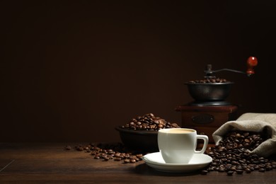 Photo of Aromatic coffee in cup, beans and grinder on wooden table against brown background, space for text
