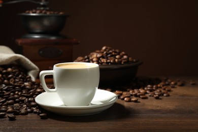Photo of Aromatic coffee in cup, beans and grinder on wooden table against brown background, closeup