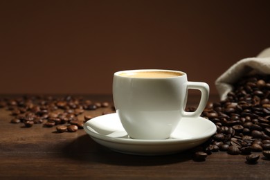 Photo of Aromatic coffee in cup and beans on wooden table against brown background, closeup