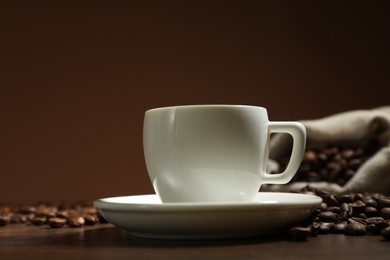 Photo of Aromatic coffee in cup and beans on wooden table against brown background, closeup