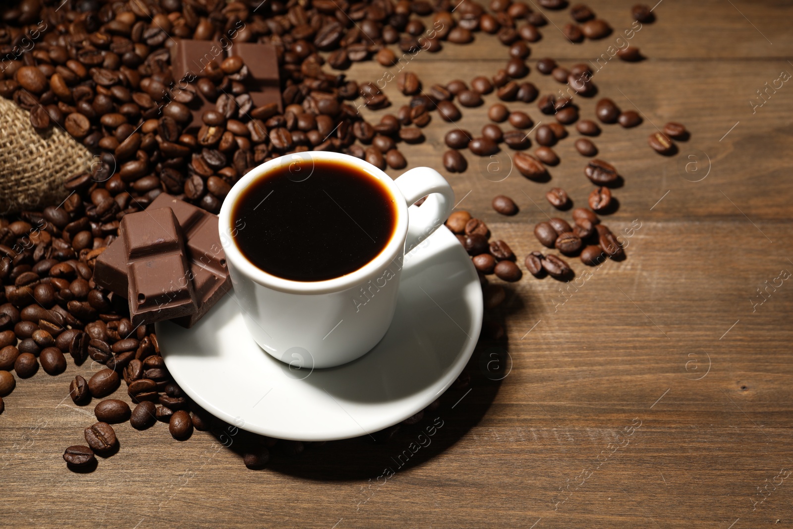 Photo of Aromatic coffee in cup, beans and pieces of chocolate on wooden table, closeup. Space for text