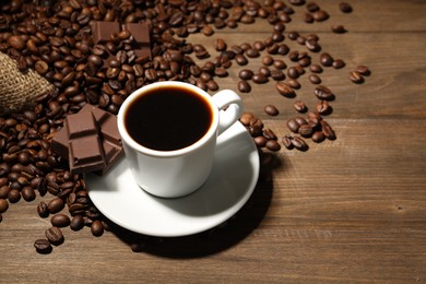 Photo of Aromatic coffee in cup, beans and pieces of chocolate on wooden table, closeup. Space for text