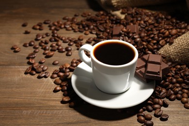 Photo of Aromatic coffee in cup, beans and pieces of chocolate on wooden table, closeup. Space for text