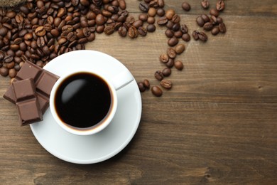 Photo of Aromatic coffee in cup, beans and pieces of chocolate on wooden table, flat lay. Space for text