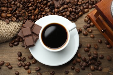 Photo of Aromatic coffee in cup, beans, pieces of chocolate and grinder on wooden table, flat lay