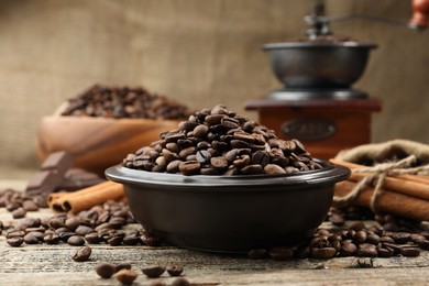 Photo of Aromatic coffee beans in bowl, cinnamon and grinder on wooden table, closeup