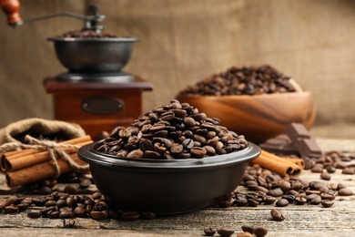 Photo of Aromatic coffee beans in bowl, cinnamon and grinder on wooden table, closeup
