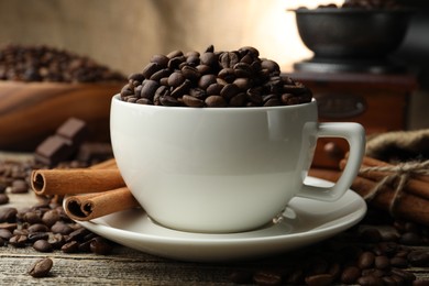 Photo of Aromatic coffee beans in cup, cinnamon and grinder on wooden table, closeup