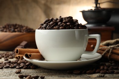 Photo of Aromatic coffee beans in cup, cinnamon and grinder on wooden table, closeup