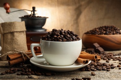 Photo of Aromatic coffee beans in cup, cinnamon and grinder on wooden table, closeup