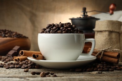 Photo of Aromatic coffee beans in cup, cinnamon and grinder on wooden table, closeup