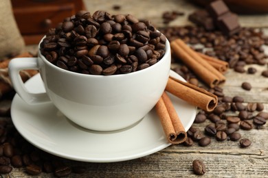 Photo of Aromatic coffee beans in cup, cinnamon and pieces of chocolate on wooden table, closeup