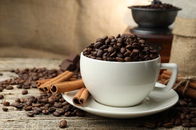 Photo of Aromatic coffee beans in cup, cinnamon and grinder on wooden table, closeup