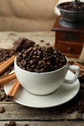 Photo of Aromatic coffee beans in cup, cinnamon and grinder on wooden table, closeup