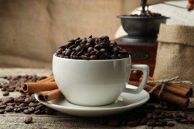 Photo of Aromatic coffee beans in cup, cinnamon and grinder on wooden table, closeup