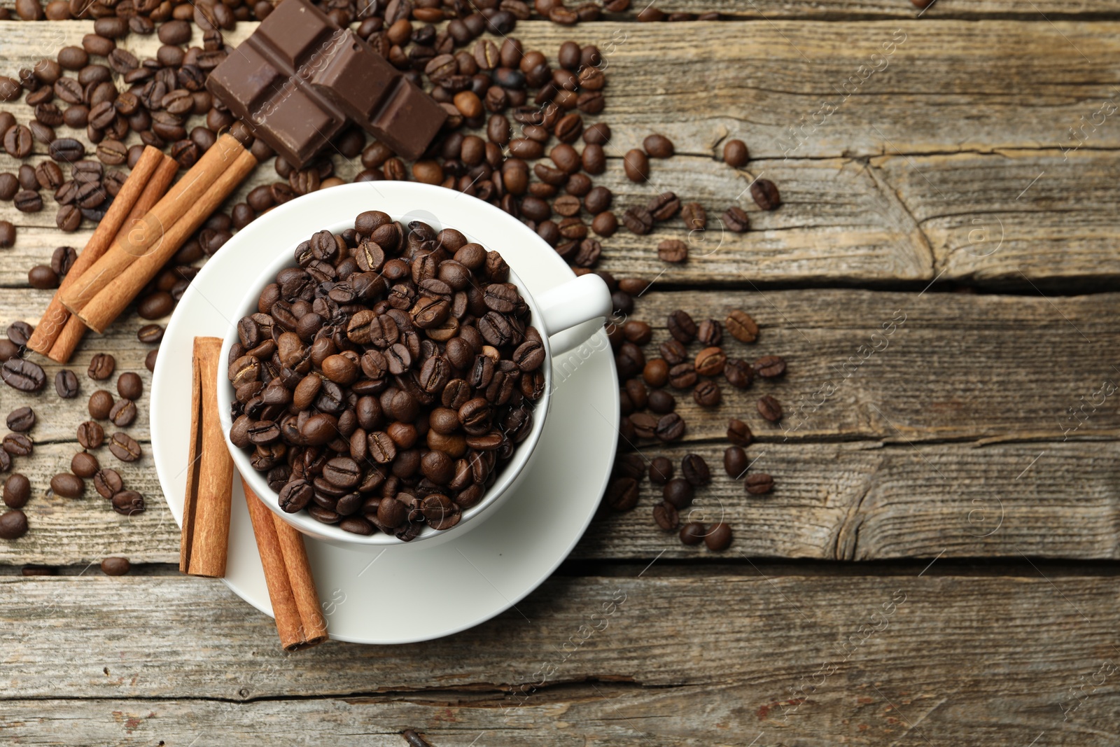 Photo of Aromatic coffee beans in cup, cinnamon and pieces of chocolate on wooden table, flat lay. Space for text