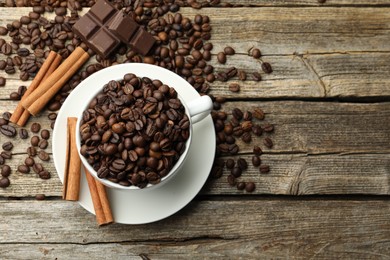 Photo of Aromatic coffee beans in cup, cinnamon and pieces of chocolate on wooden table, flat lay. Space for text