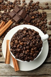 Photo of Aromatic coffee beans in cup, cinnamon and pieces of chocolate on wooden table, flat lay