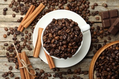 Photo of Aromatic coffee beans in cup, cinnamon and pieces of chocolate on wooden table, flat lay
