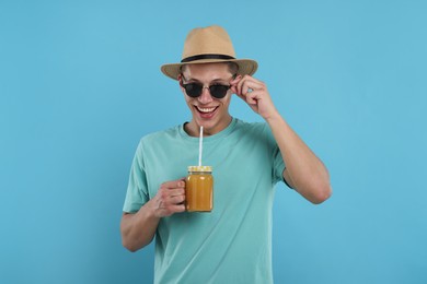 Photo of Happy man in sunglasses and hat with mason jar of orange juice on light blue background. Refreshing drink