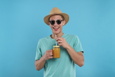 Photo of Happy man in sunglasses and hat with mason jar of orange juice on light blue background. Refreshing drink