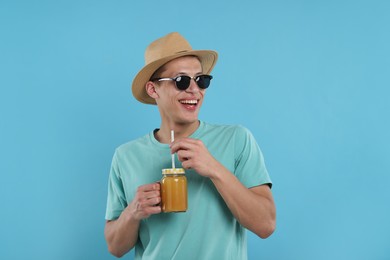 Photo of Happy man in sunglasses and hat with mason jar of orange juice on light blue background. Refreshing drink