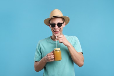 Photo of Happy man in sunglasses and hat with mason jar of orange juice on light blue background. Refreshing drink