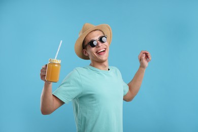 Photo of Happy man in sunglasses and hat with mason jar of orange juice on light blue background. Refreshing drink