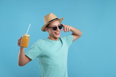 Photo of Happy man in sunglasses and hat with mason jar of orange juice on light blue background. Refreshing drink