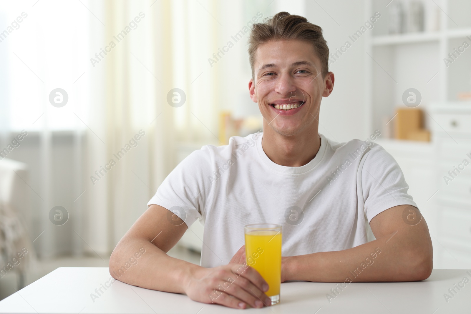 Smiling man with glass of orange juice at white table indoors. Refreshing drink Photo of Smiling man with glass of orange juice at white table indoors. Refreshing drink