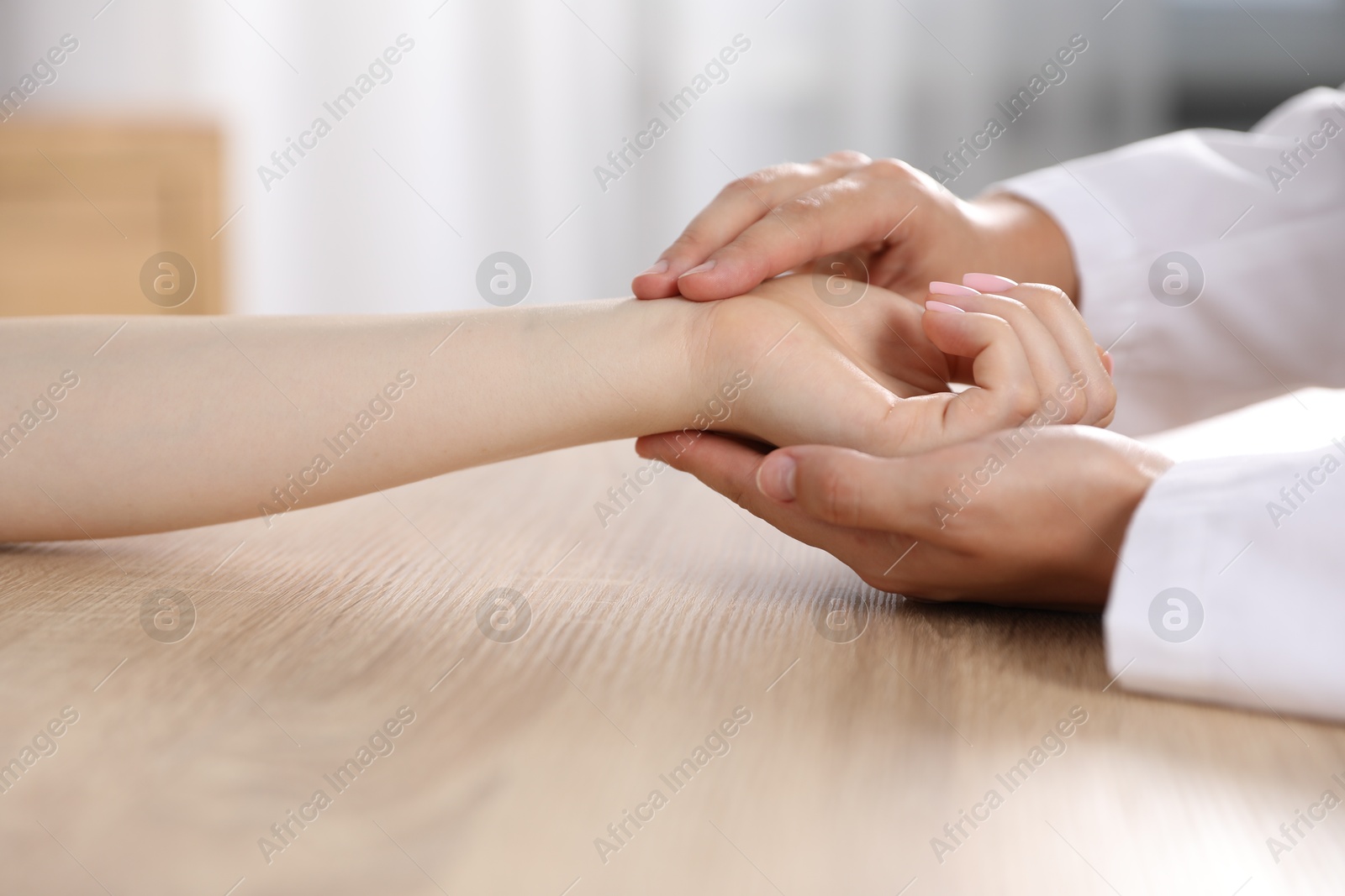 Photo of Doctor checking woman's pulse with fingers at wooden table indoors, closeup
