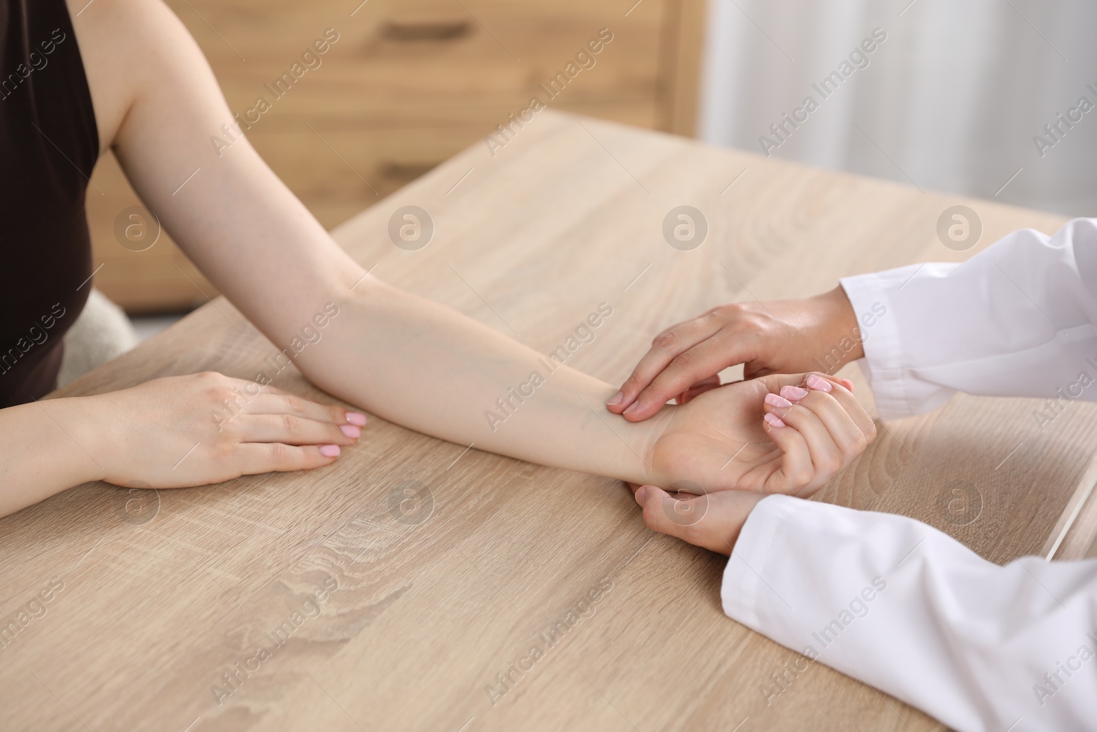 Photo of Doctor checking woman's pulse with fingers at wooden table indoors, closeup