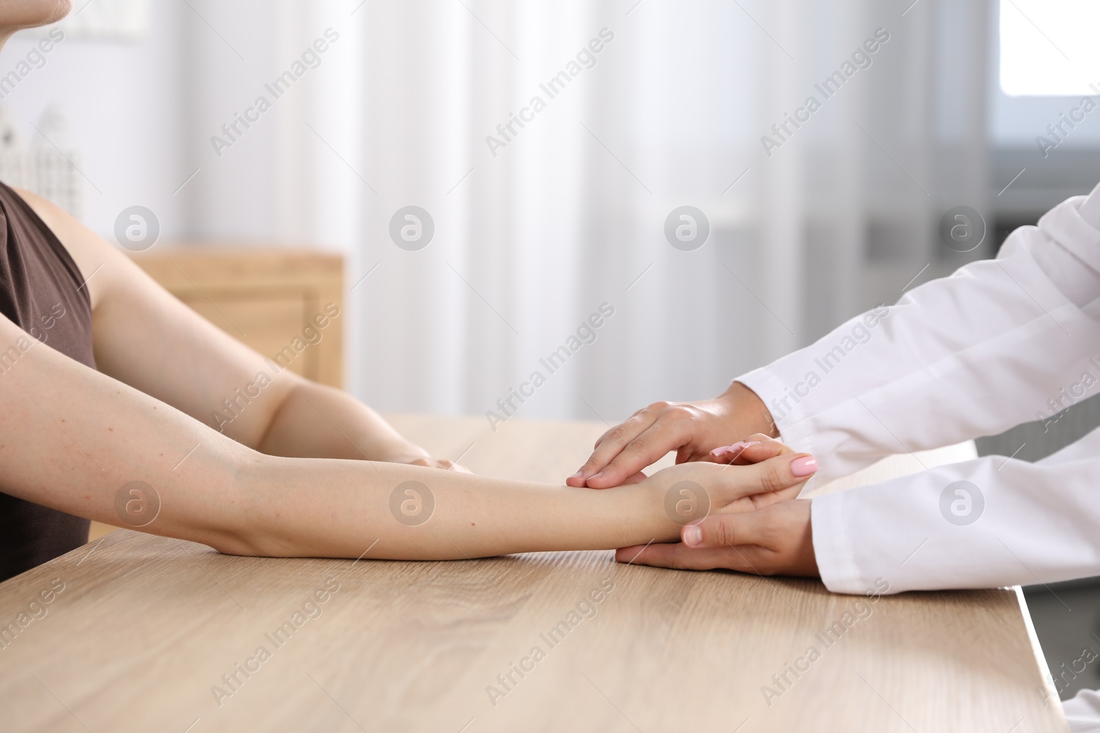 Photo of Doctor checking woman's pulse with fingers at wooden table indoors, closeup