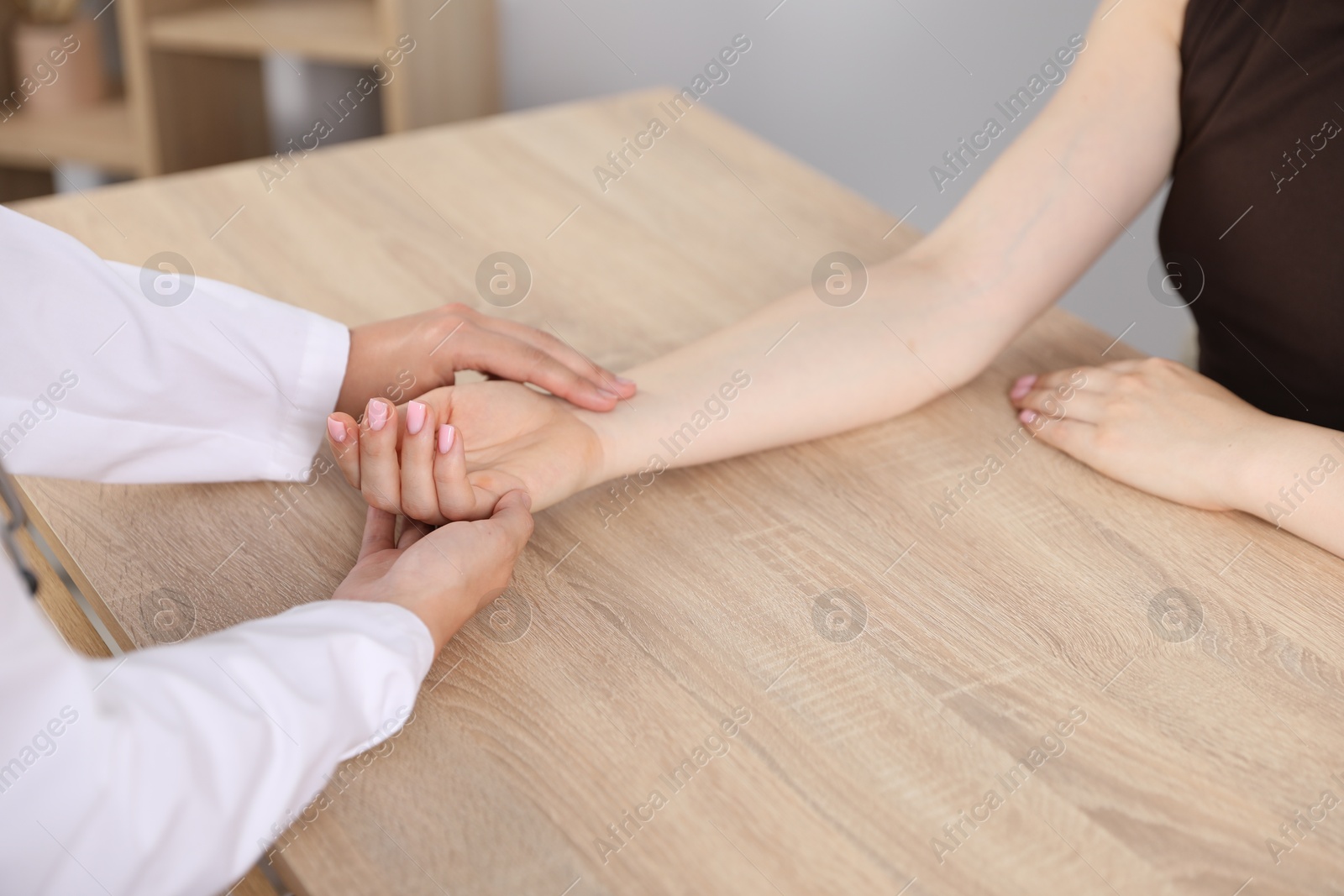 Photo of Doctor checking woman's pulse with fingers at wooden table indoors, closeup