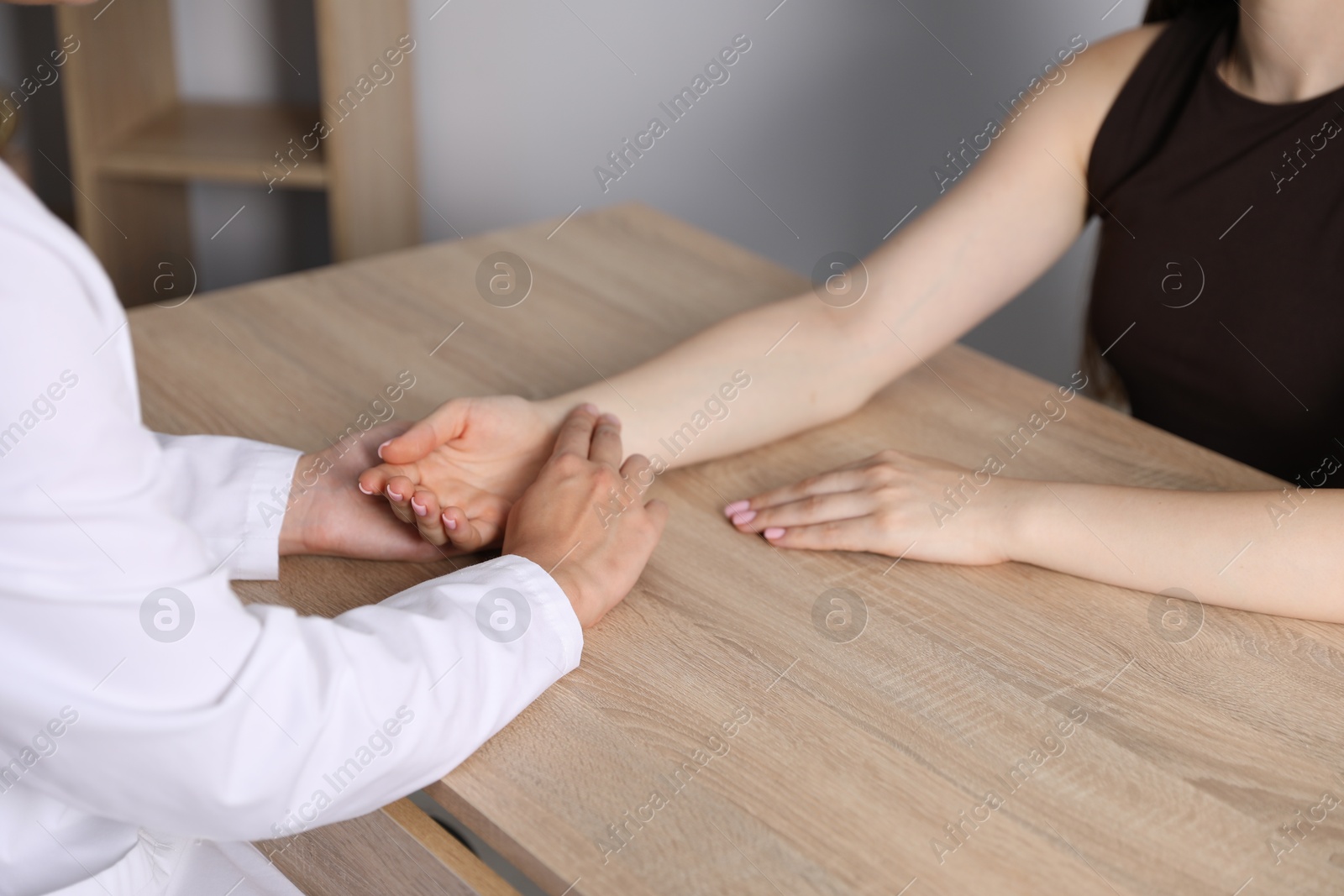 Photo of Doctor checking woman's pulse with fingers at wooden table indoors, closeup