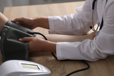 Photo of Doctor checking woman's heart rate and blood pressure via sphygmomanometer at wooden table indoors, closeup