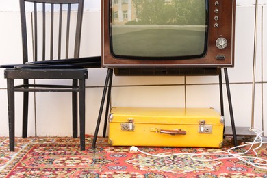 Photo of Retro TV on stand, suitcase and black chair near white wall. Used stuff