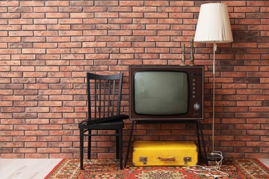 Photo of Retro TV on stand, suitcase, lamp and black chair near red brick wall, space for text. Used stuff