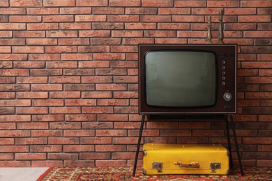 Photo of Retro TV on stand, suitcase and carpet near red brick wall, space for text. Used stuff