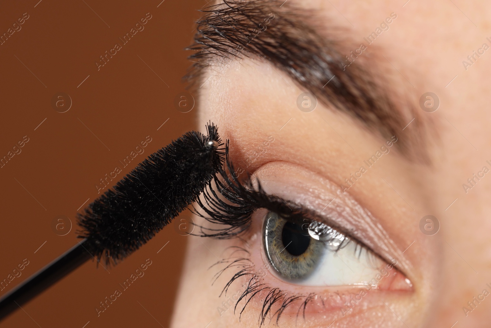Photo of Woman applying mascara onto her eyelashes on brown background, closeup